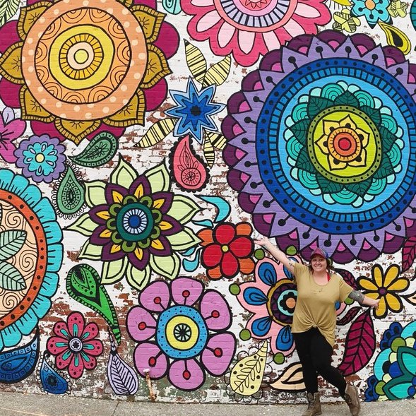 Person posing in front of a colorful mural of flowers and mandalas.