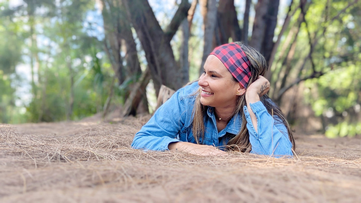 A woman lying on dry grass in a wooded outdoor setting, wearing a blue denim shirt and a red and black plaid headband.