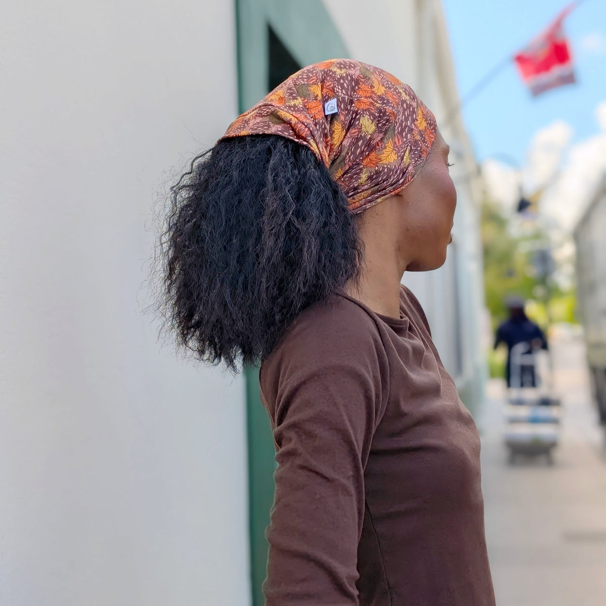 A woman in profile wearing a brown long-sleeve shirt and a patterned orange, yellow, and red headwrap headscarf, standing outdoors next to a white building.