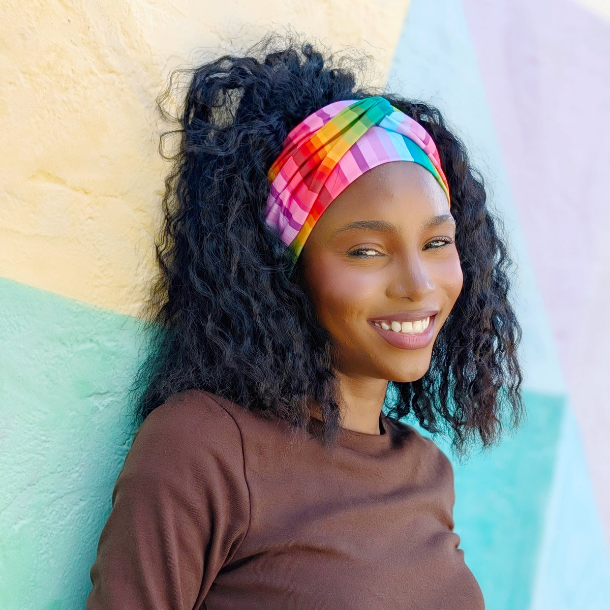 A smiling woman with curly black hair wears a vibrant rainbow-striped plaid headband and a brown long-sleeve top, leaning against a pastel geometric wall.