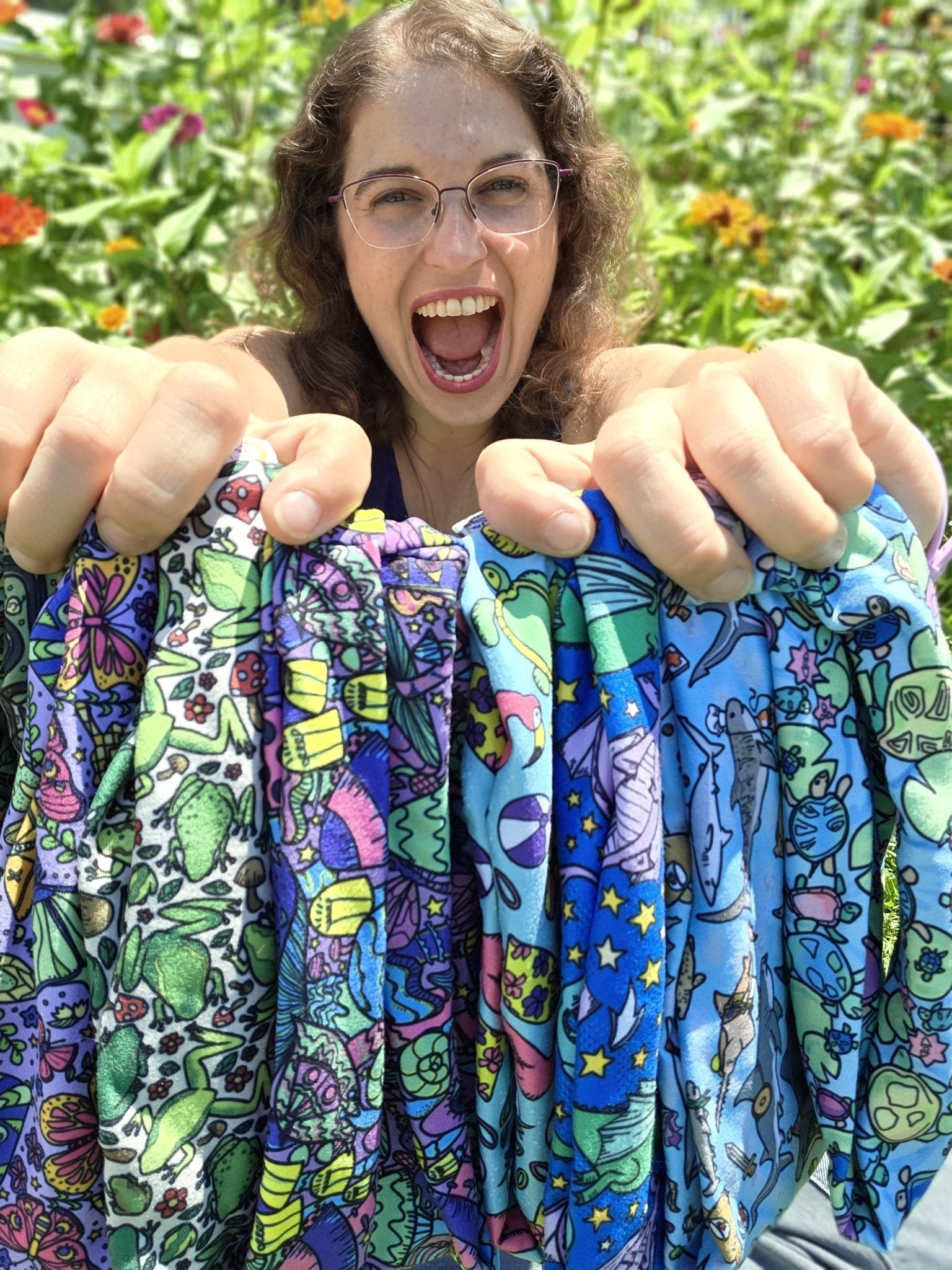 A smiling woman outdoors in a garden holds up several colorful patterned fabric pieces with cartoon space-themed designs like planets, stars, rockets, and aliens.