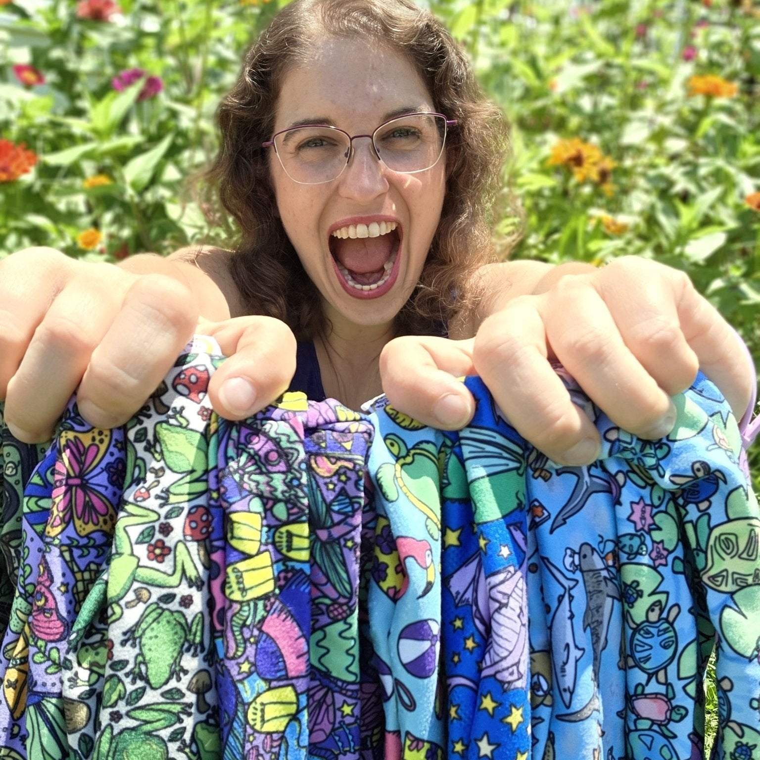 A smiling woman outdoors in a garden holds up several colorful patterned fabric pieces with cartoon space-themed designs like planets, stars, rockets, and aliens.