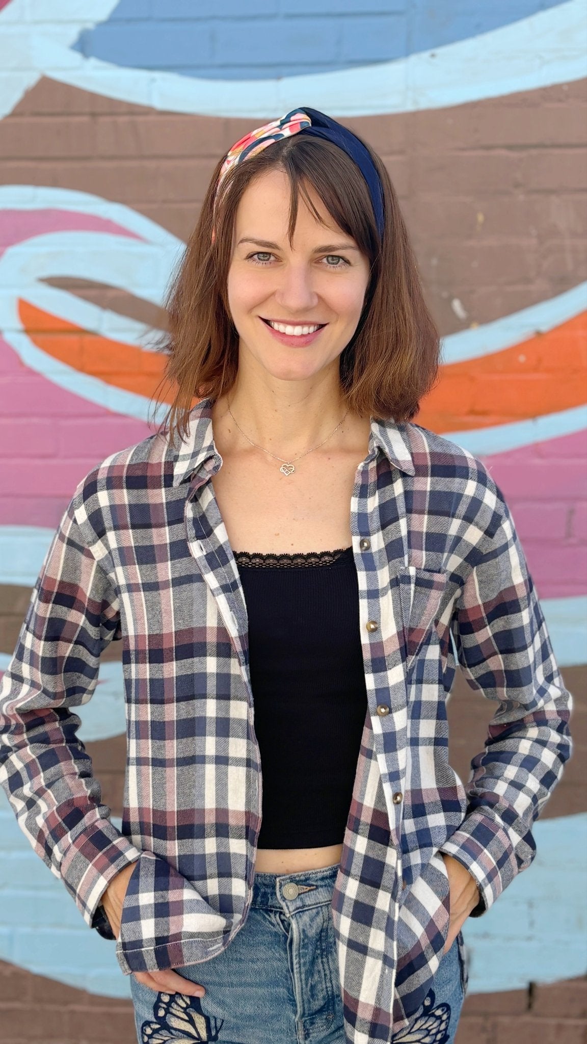 A woman smiling in front of a colorful graffiti wall, wearing a navy, red, and white plaid button-up shirt over a black crop top and blue jeans.