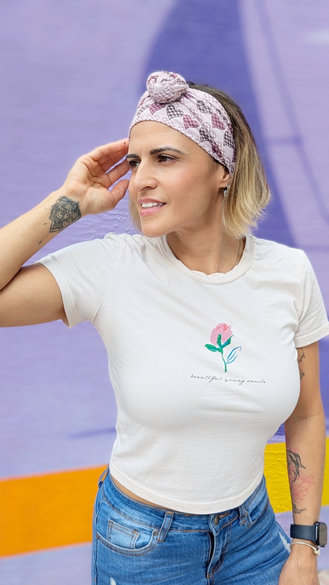 A woman wearing a white graphic t-shirt with a pink rose design and blue jeans, paired with a pink and white patterned headband, posing against a purple and orange background.