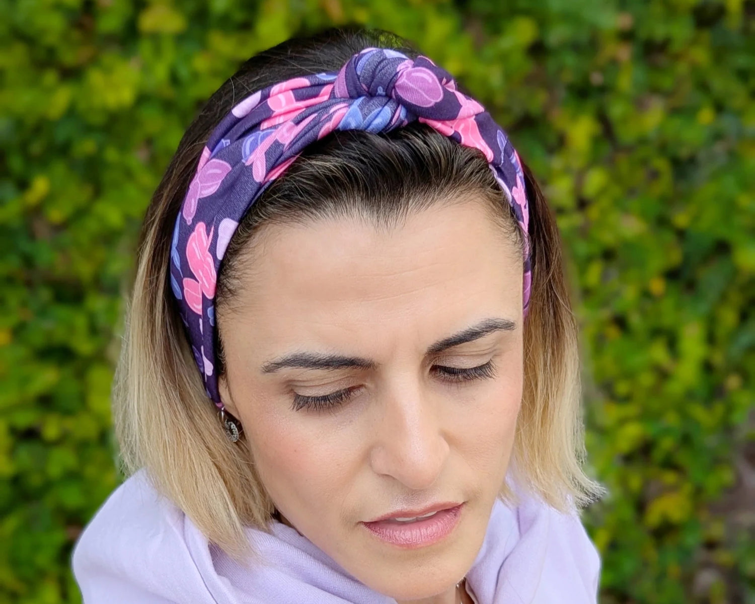 Woman wearing a light purple hoodie and colorful headband with greenery in the background