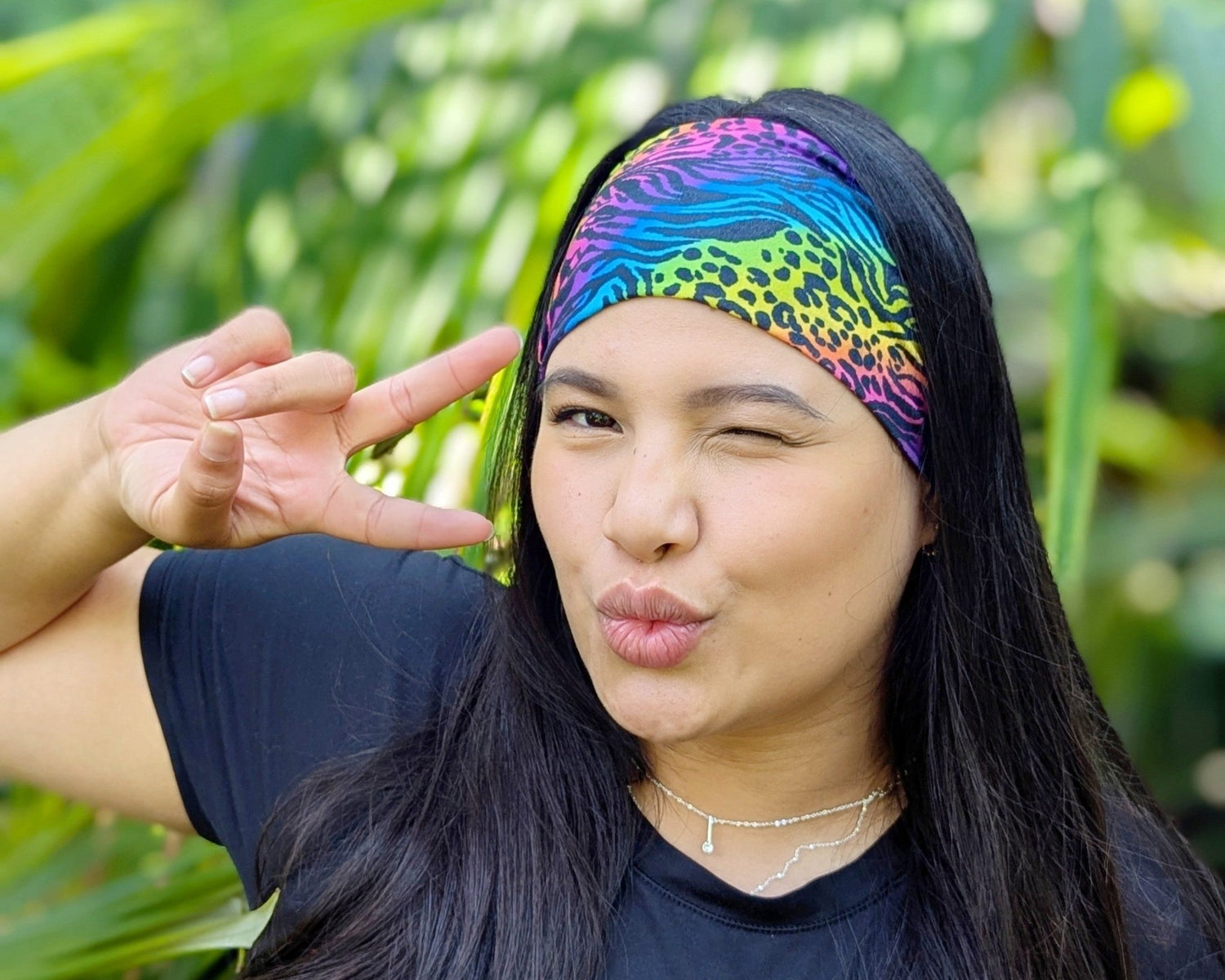 Person wearing a colorful headband with a leopard print pattern, making a peace sign in front of green foliage.