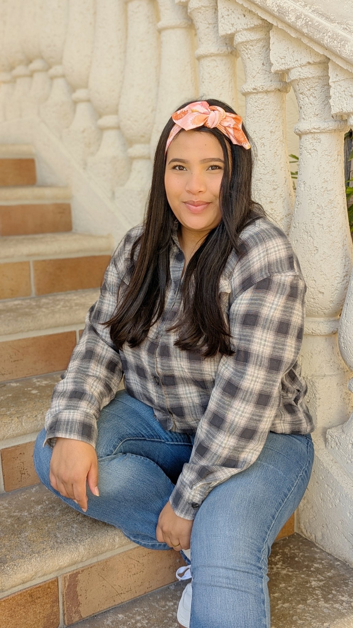 A woman sitting on outdoor stone steps wearing a gray and white plaid flannel shirt, blue jeans, and a pink bow headband.