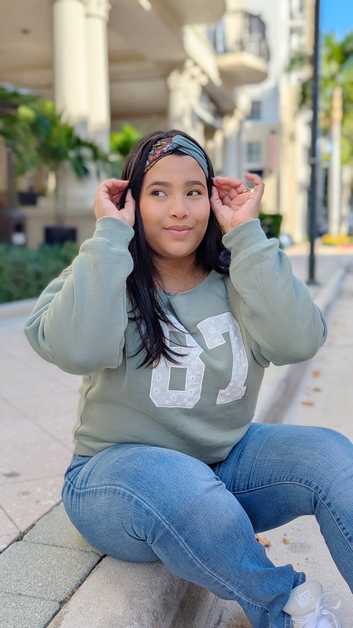 A woman sitting outdoors on a curb wearing a sage green crewneck sweatshirt with a white '87' graphic, blue jeans, and a patterned headband.