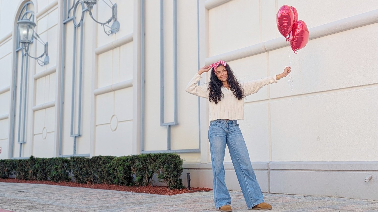 Woman holding pink balloons in an urban setting with a light-colored building.