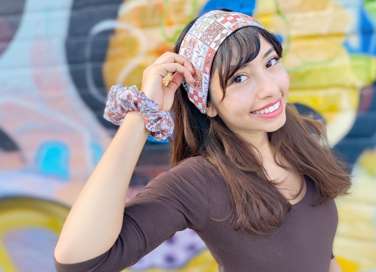 A woman wearing a patterned headband and matching scrunchie, posing in front of a colorful graffiti wall.
