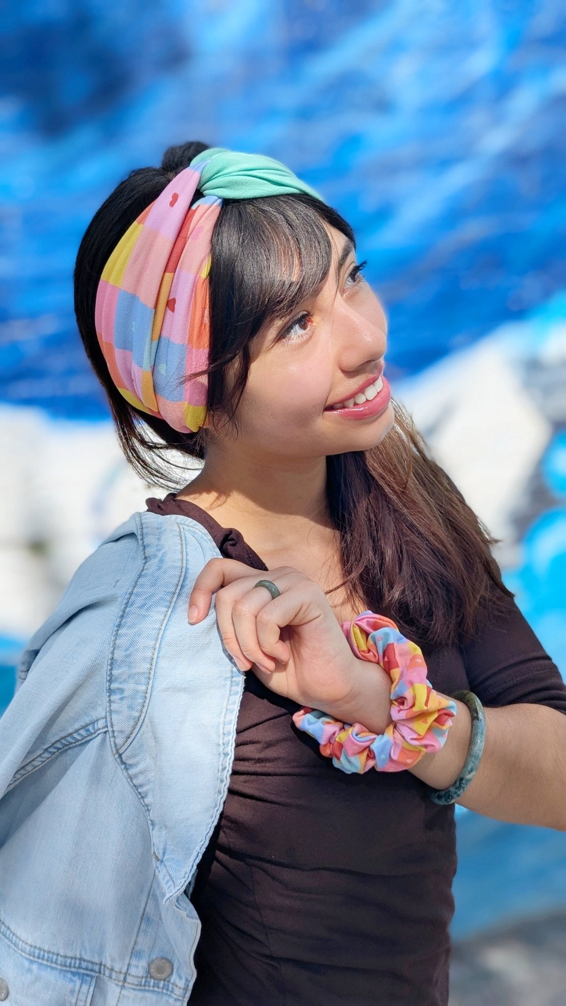 A woman outdoors wearing a pastel multicolor patterned headband and matching scrunchie, with a light blue denim jacket draped over her shoulder and a dark brown top.