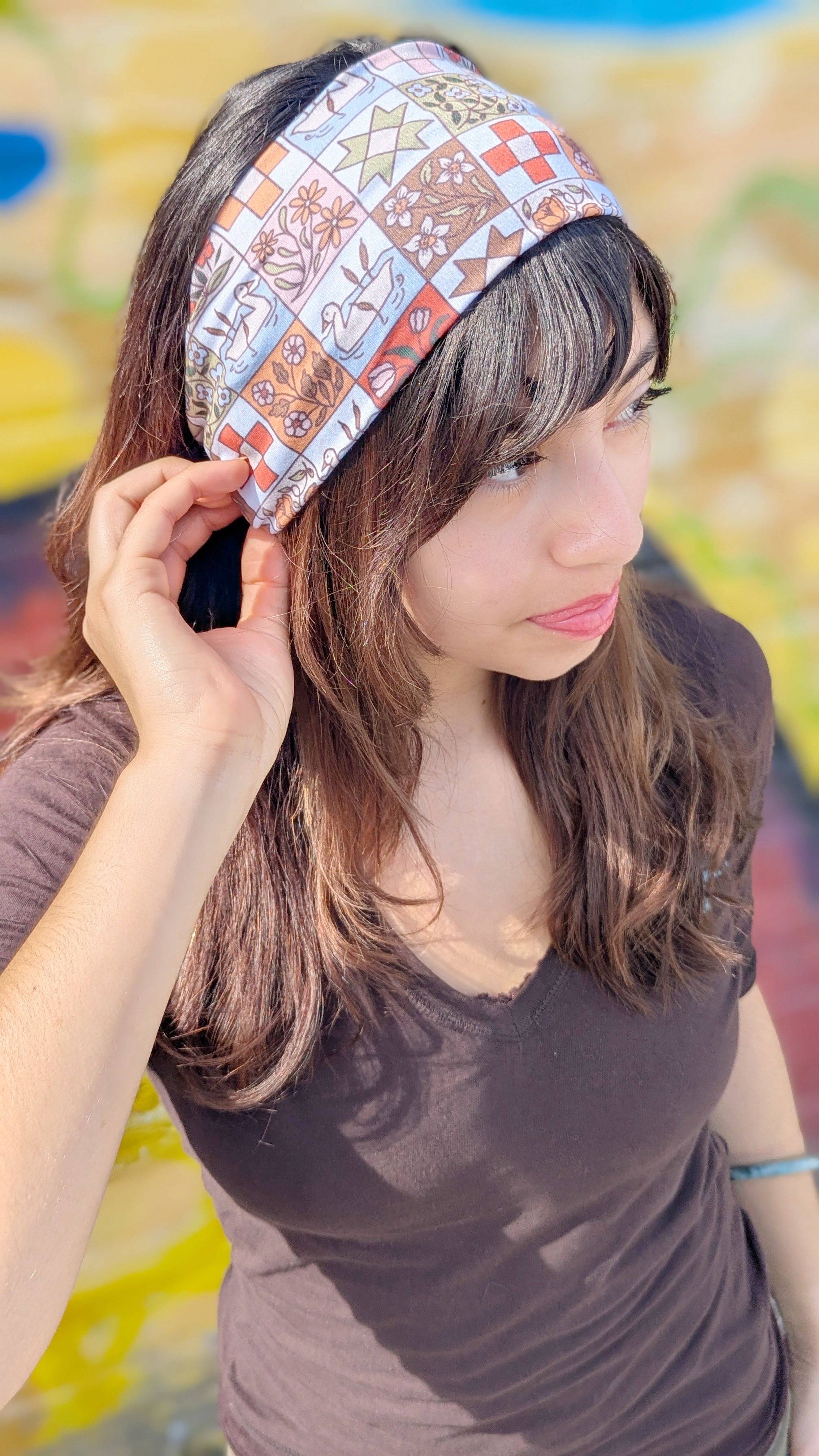 A woman wearing a patterned headband and a dark brown v-neck t-shirt, posing outdoors in front of a colorful, blurred background.