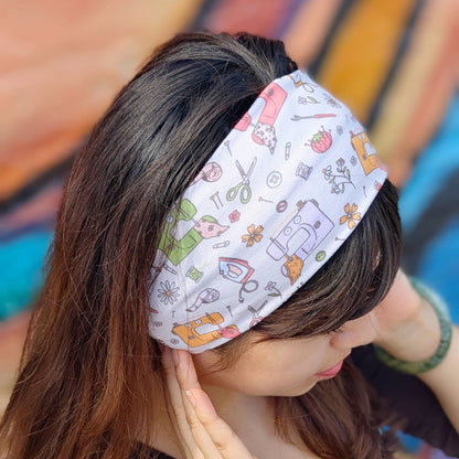 A woman wearing a white fabric headband with a colorful cartoon pattern of scissors, paintbrushes, and paint splatters, paired with a brown long-sleeve top.