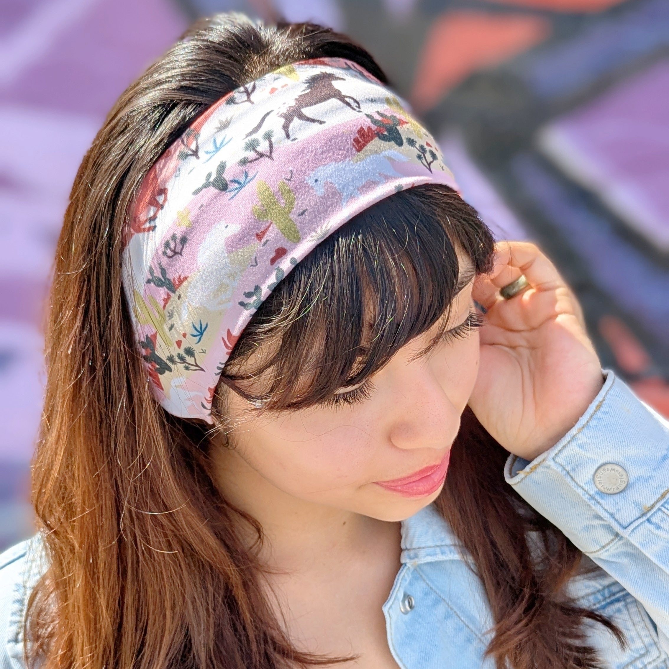 A woman wearing a light pink floral patterned headband, a light blue denim jacket, and a brown top, posing outdoors with a blurred graffiti wall in the background.