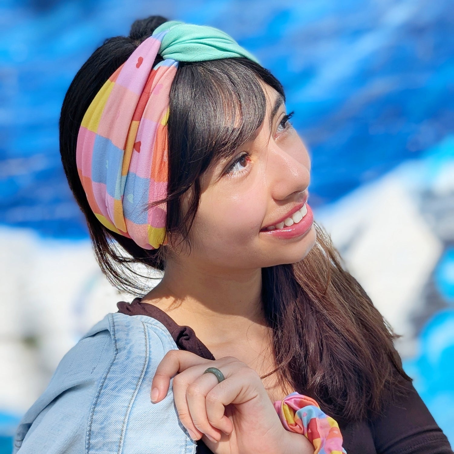 A woman outdoors wearing a pastel multicolor patterned headband and matching scrunchie, with a light blue denim jacket draped over her shoulder and a dark brown top.