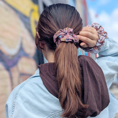 A person viewed from behind wearing a light blue denim jacket and a brown hoodie, adjusting a pink and blue patterned scrunchie in their brown ponytail outdoors against a graffiti wall.