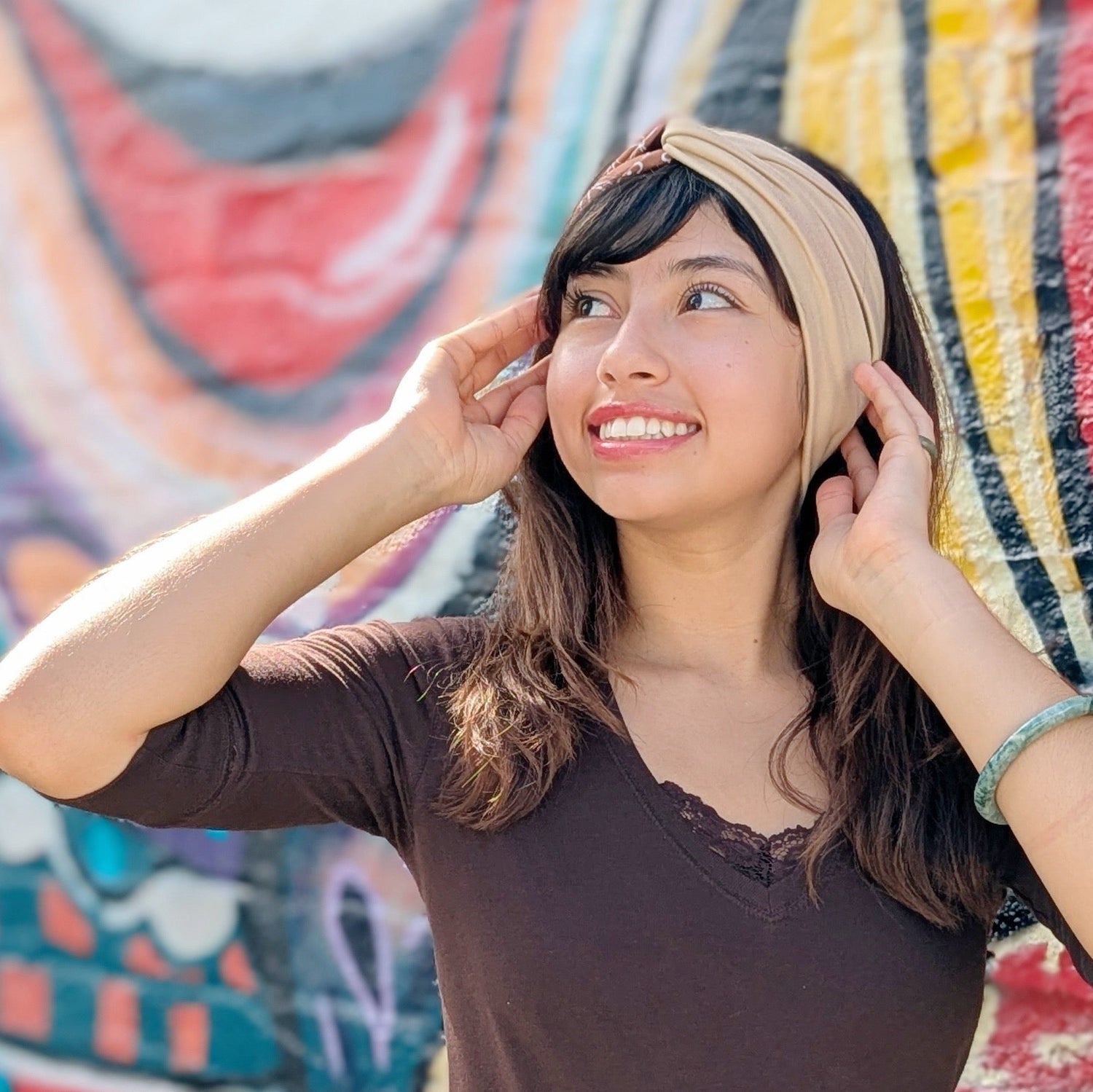 A woman wearing a beige fabric headband and a dark brown v-neck top, posing in front of a colorful graffiti wall.