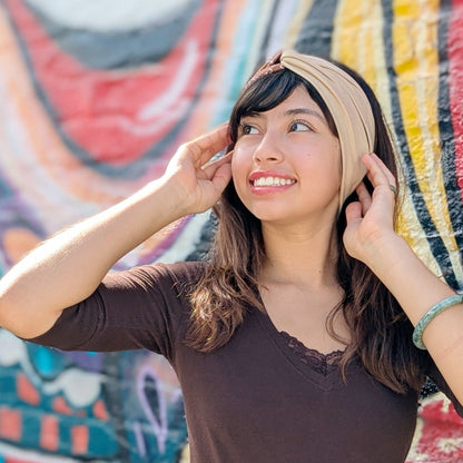 A woman wearing a beige fabric headband and a dark brown v-neck top, posing in front of a colorful graffiti wall.