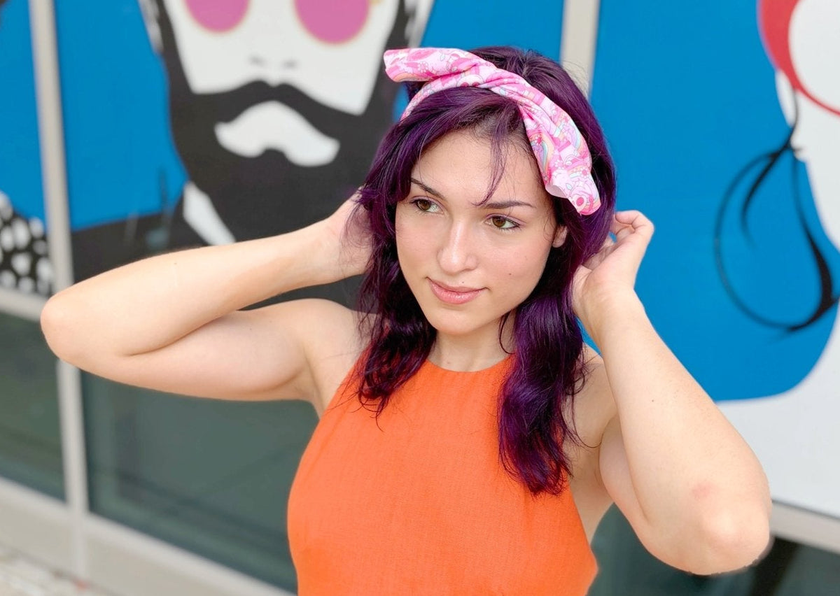 A woman with purple hair wearing a pink patterned headband and an orange top, posing in front of a blue and white graphic mural.