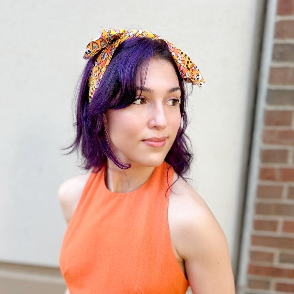 A woman with purple hair wearing a bright orange sleeveless dress and a patterned headband, holding a small orange pumpkin. She is standing outdoors against a beige wall and brick column.