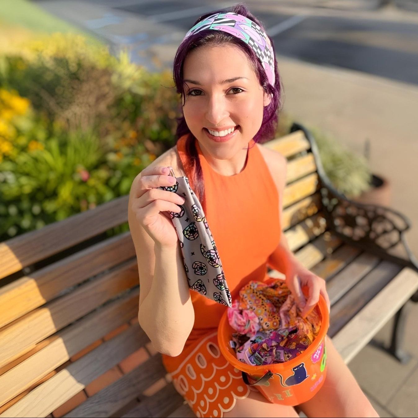 A woman with purple hair sits on a wooden park bench, smiling and holding a patterned phone case and an orange bucket filled with colorful items. She is wearing an orange sleeveless top and a multicolor patterned headband in an outdoor setting with a blurred background.