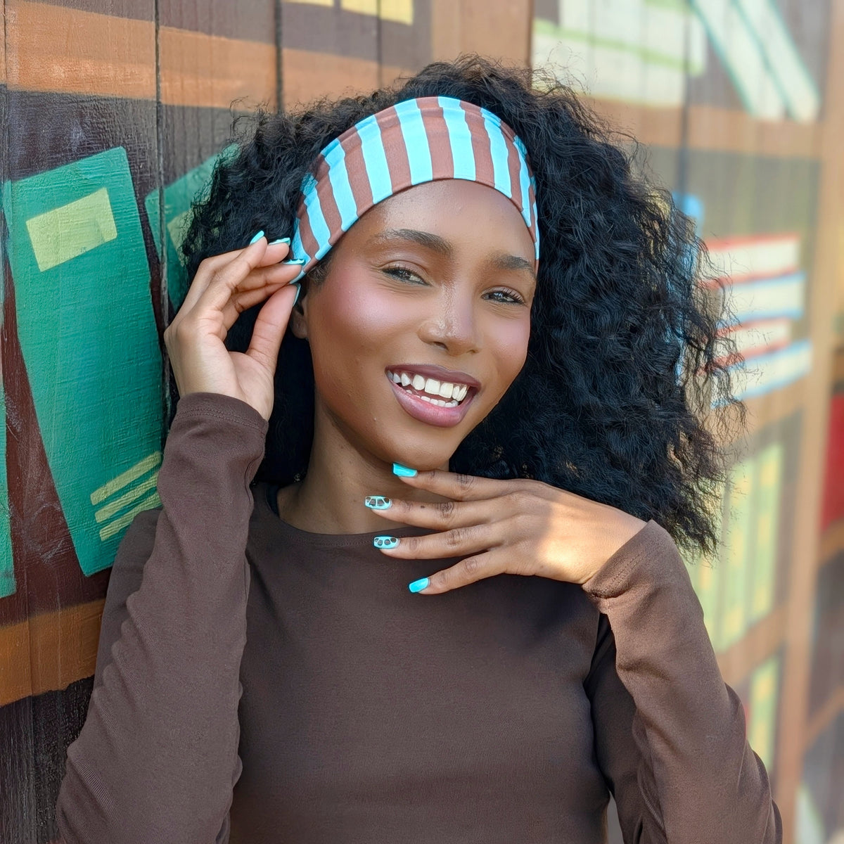 A smiling woman with curly black hair wears a brown long-sleeve top and a light blue and red striped headband, posing in front of a colorful wooden wall.