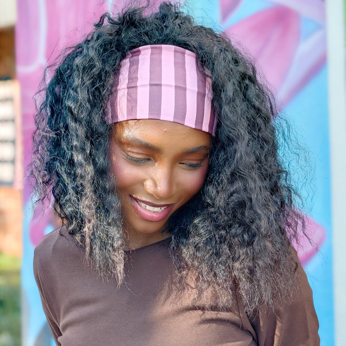 A woman with long, curly black hair wearing a pink and purple striped headband and a brown top, looking down with a smile in front of a pink and blue floral background.