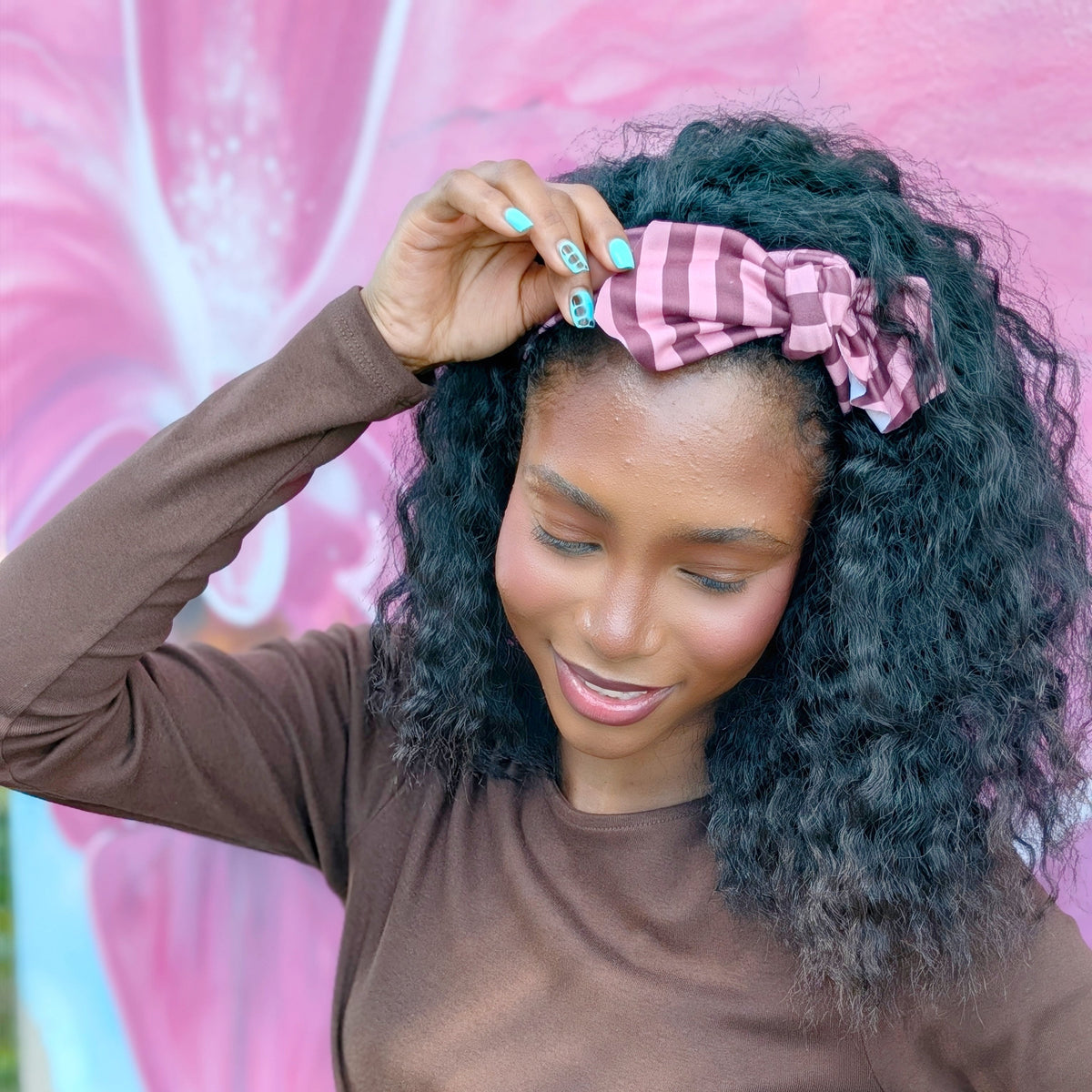 A woman with curly black hair wearing a pink and white gingham bow headband, posing in front of a pink floral mural.