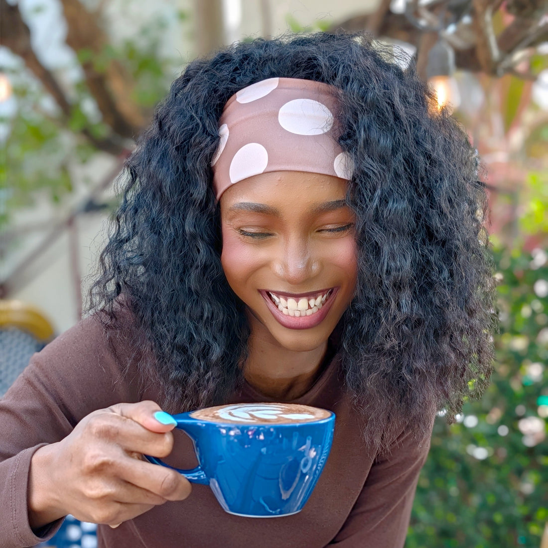 A woman with curly black hair wearing a pink polka-dot headband and a brown long-sleeve top, smiling while holding a blue mug with latte art in an outdoor cafe setting.