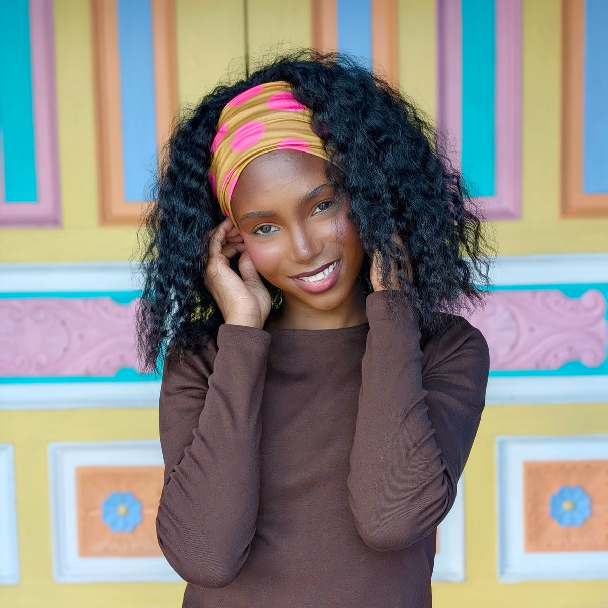A woman with curly black hair wearing a brown long-sleeve top and a pink and yellow striped headband, posing in front of a colorful yellow, pink, and blue patterned wall.
