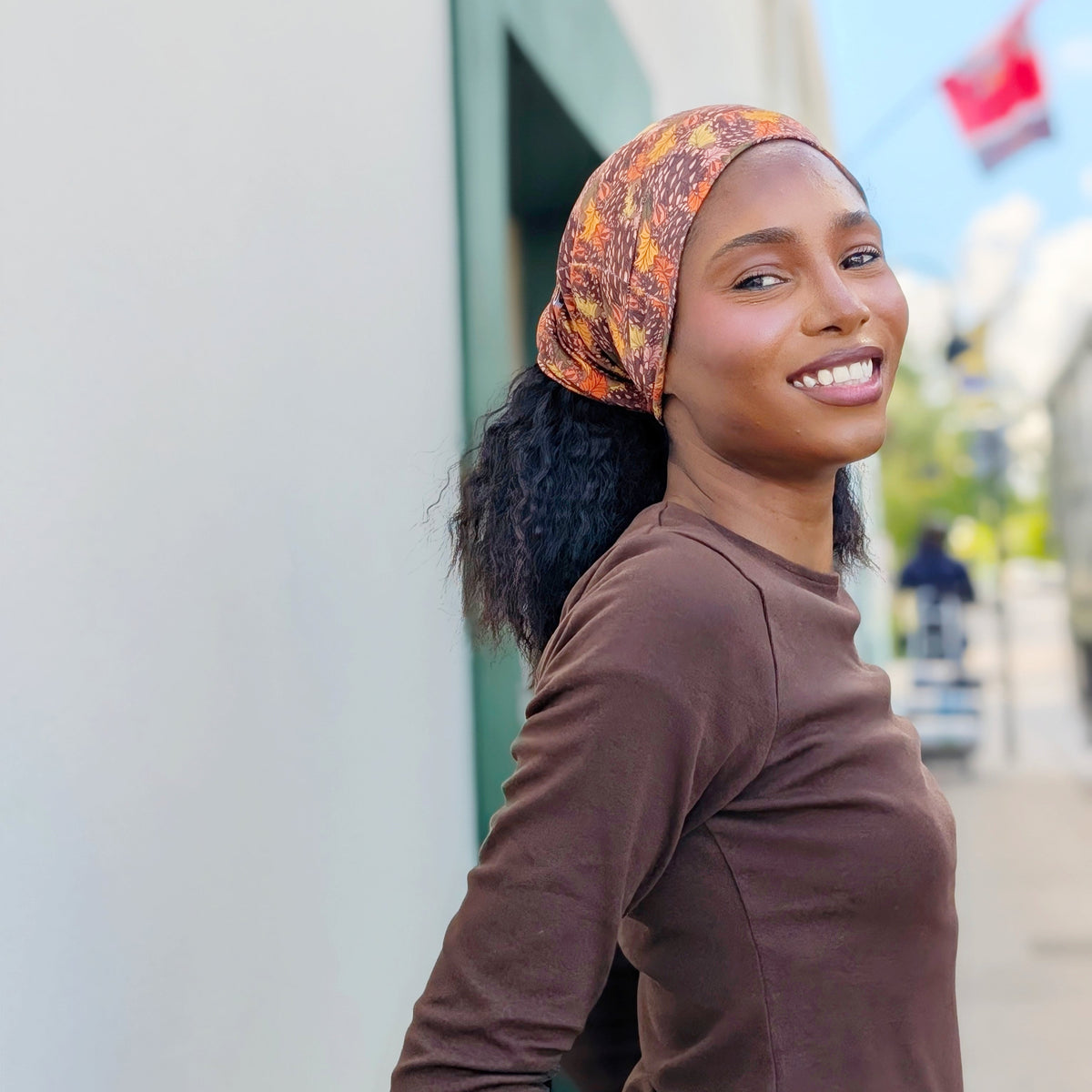 A woman wearing a brown long-sleeve top and a patterned brown, orange, and yellow headwrap headscarf, smiling and looking over her shoulder in an outdoor street setting.