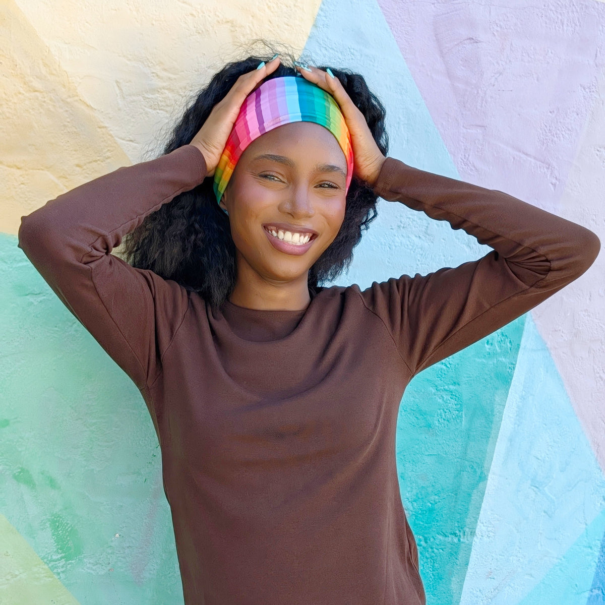 A smiling woman with curly black hair wears a brown long-sleeve top and a rainbow-striped headband, posing against a pastel geometric wall.