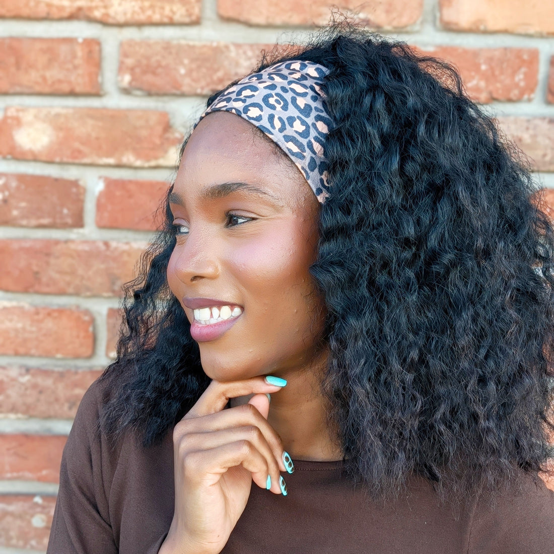 A woman with curly black hair wearing a blue and white patterned headband and a brown long-sleeve top, posing in front of a red brick wall.
