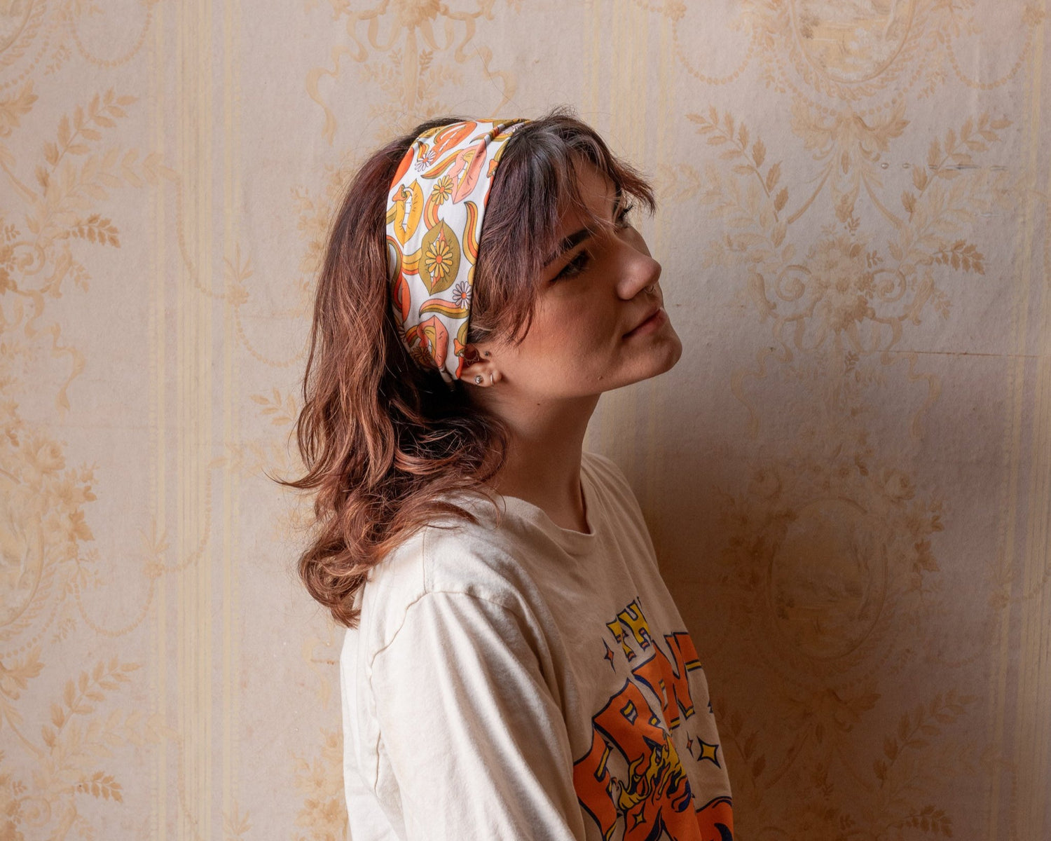 Woman wearing a patterned headband and a white shirt with orange text against a beige wall.