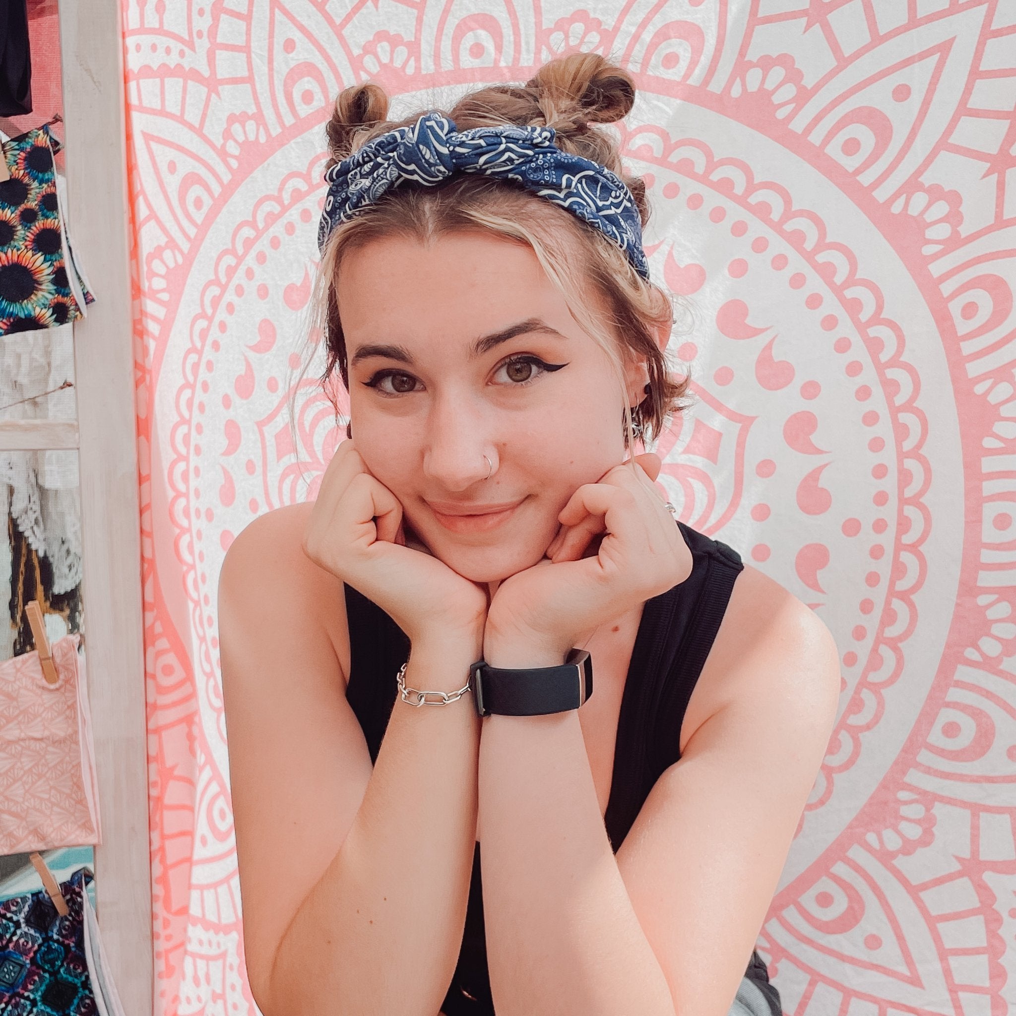 A young woman with blonde hair styled in two buns wears a blue and white patterned bandana headband, a black sleeveless top, and a black smartwatch. She is posing in front of a pink and white mandala tapestry.
