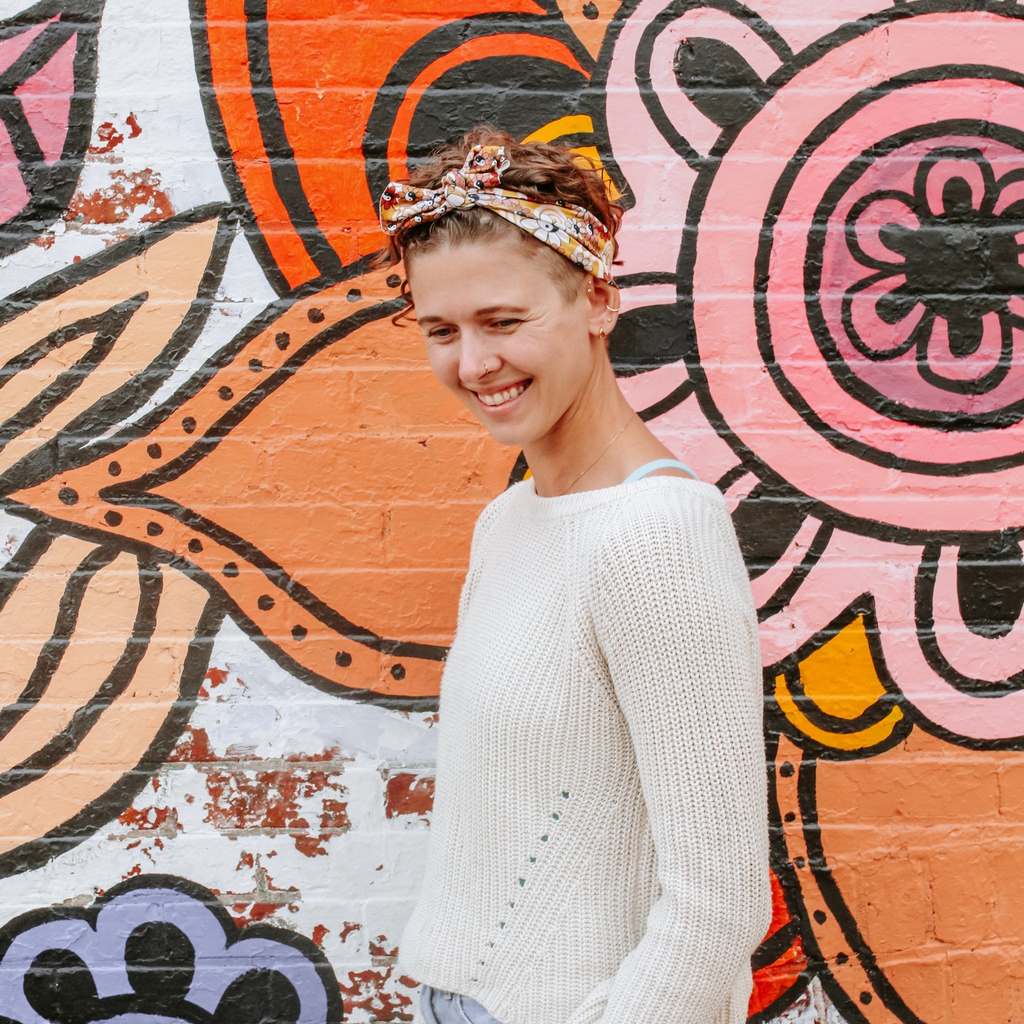 A woman wearing a cream-colored knit sweater and a patterned headband stands in front of a colorful graffiti brick wall.