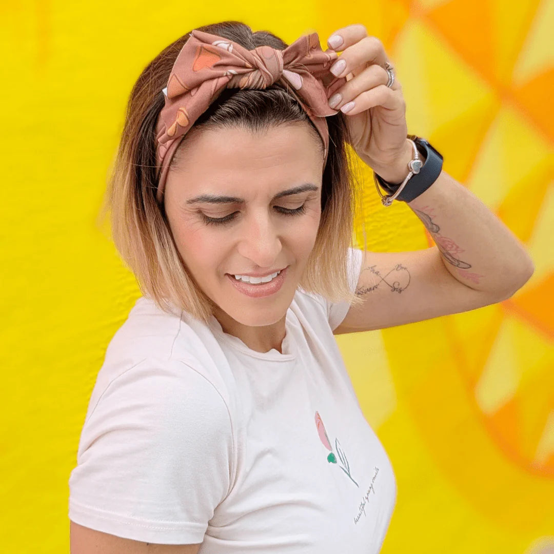 A woman with blonde-highlighted hair wearing a dusty rose patterned fabric bow headband and a white t-shirt, posing against a bright yellow background.