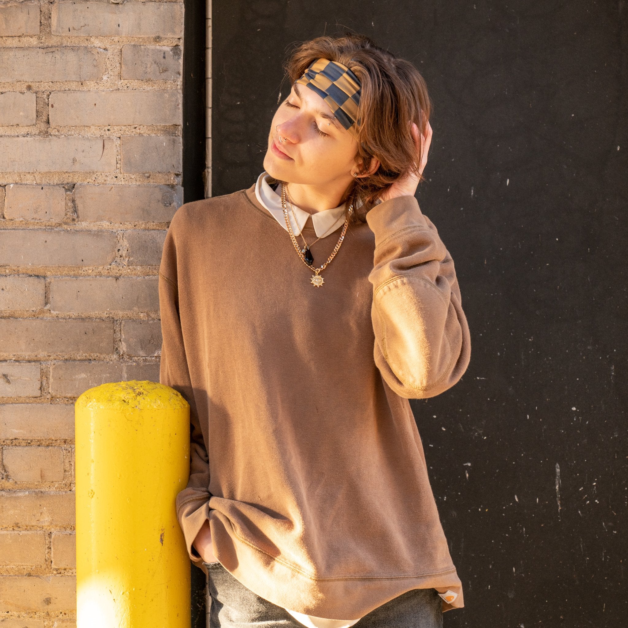 A person wearing a brown crewneck sweatshirt over a white collared shirt, standing outdoors next to a yellow post against a brick wall and dark door.