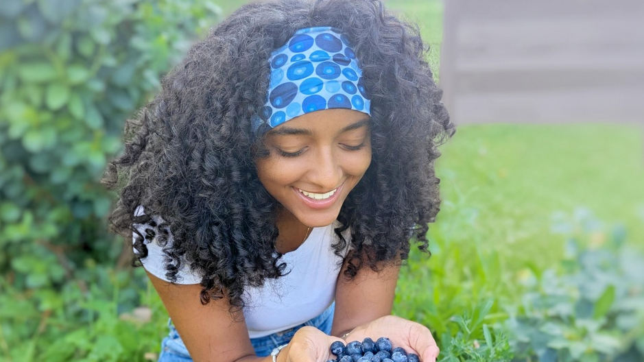 Woman with curly hair wearing a blue headband holding blueberries outdoors.