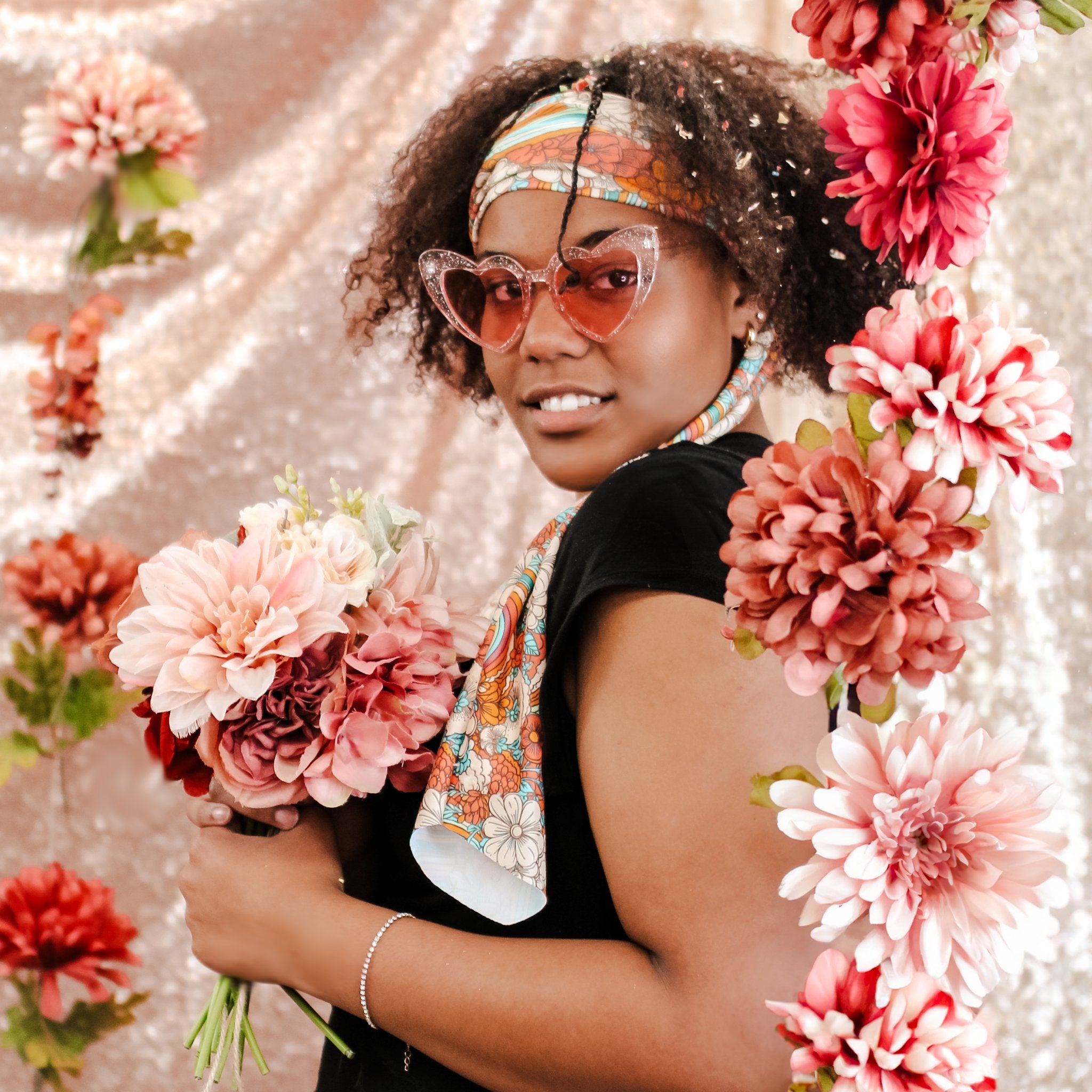 A woman wearing heart-shaped sunglasses, a patterned headband, and a colorful scarf, holding a bouquet of pink and red flowers against a shimmering pink backdrop decorated with large artificial flowers.