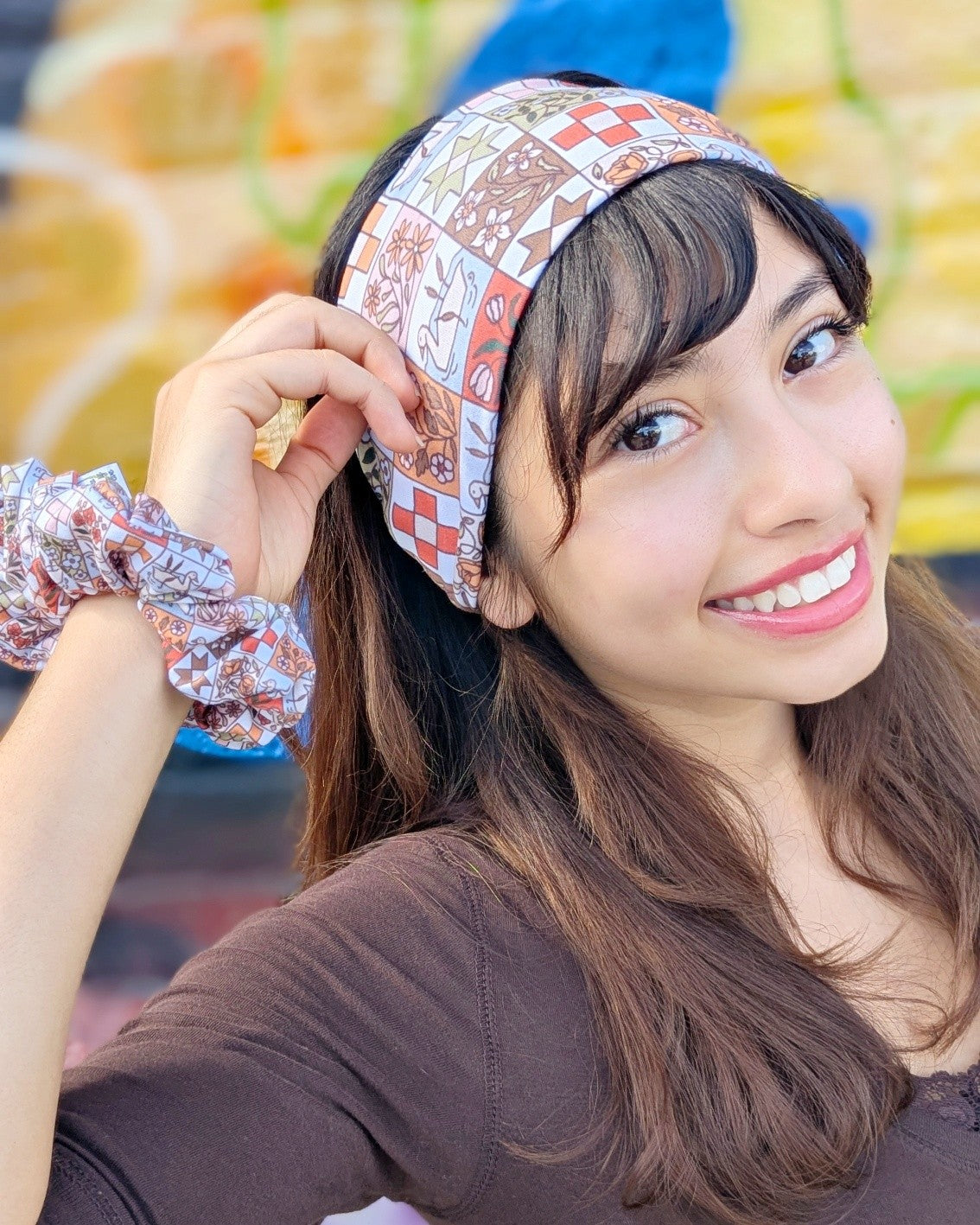 A smiling woman wearing a patterned headband and a matching patterned scrunchie, photographed in a colorful real-world setting.