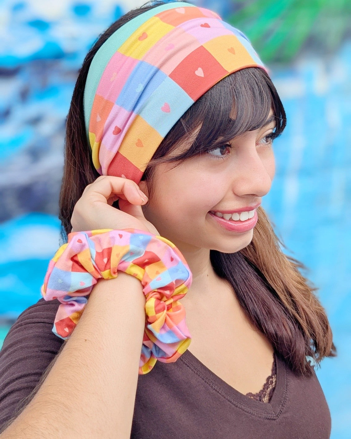 A woman wearing a colorful patchwork scrunchie headband and matching scrunchie, smiling and posing against a blurred blue background.