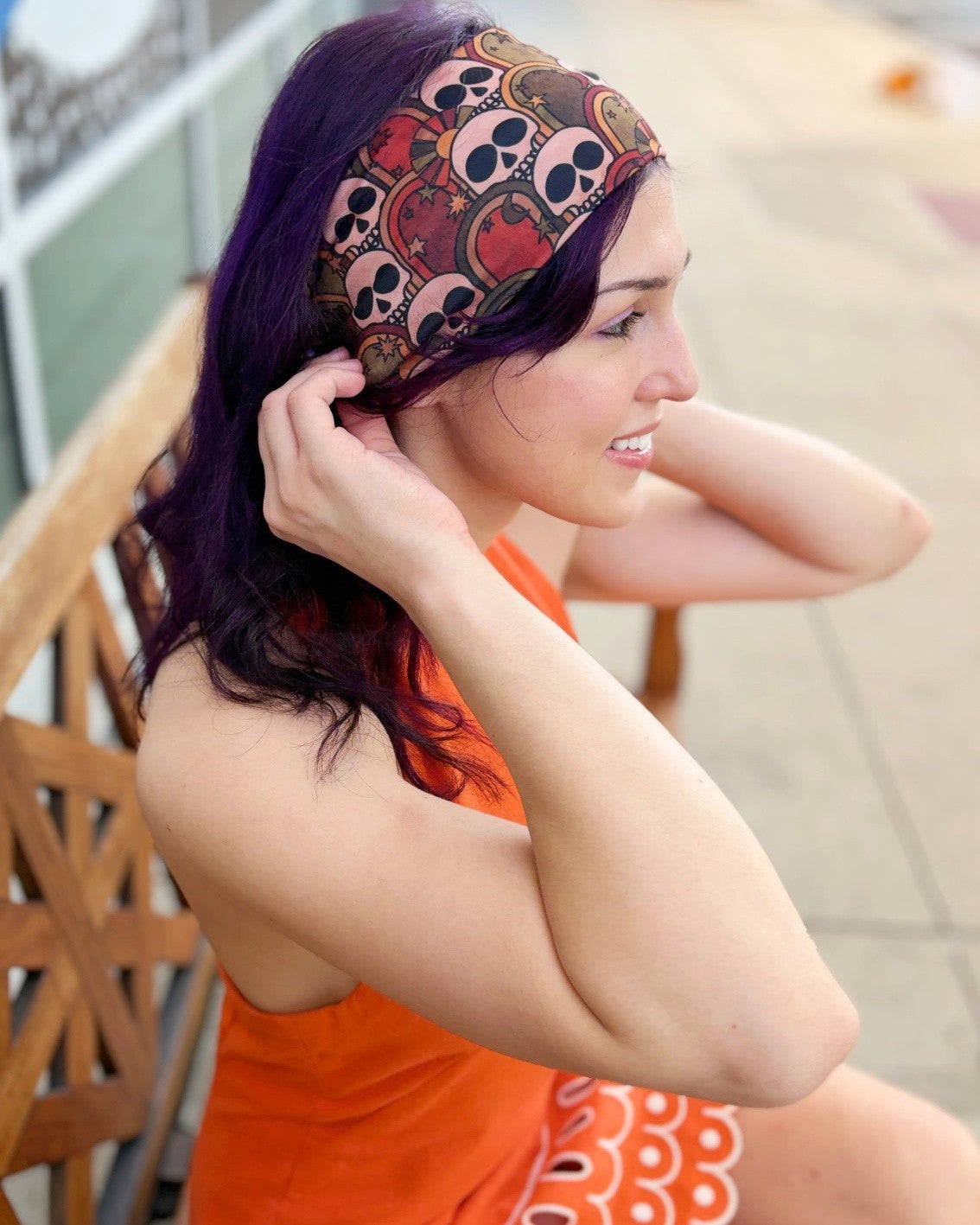 A woman with dark purple hair wearing a brown patterned skull and crossbones headband and an orange dress, sitting on a wooden bench in an outdoor setting.